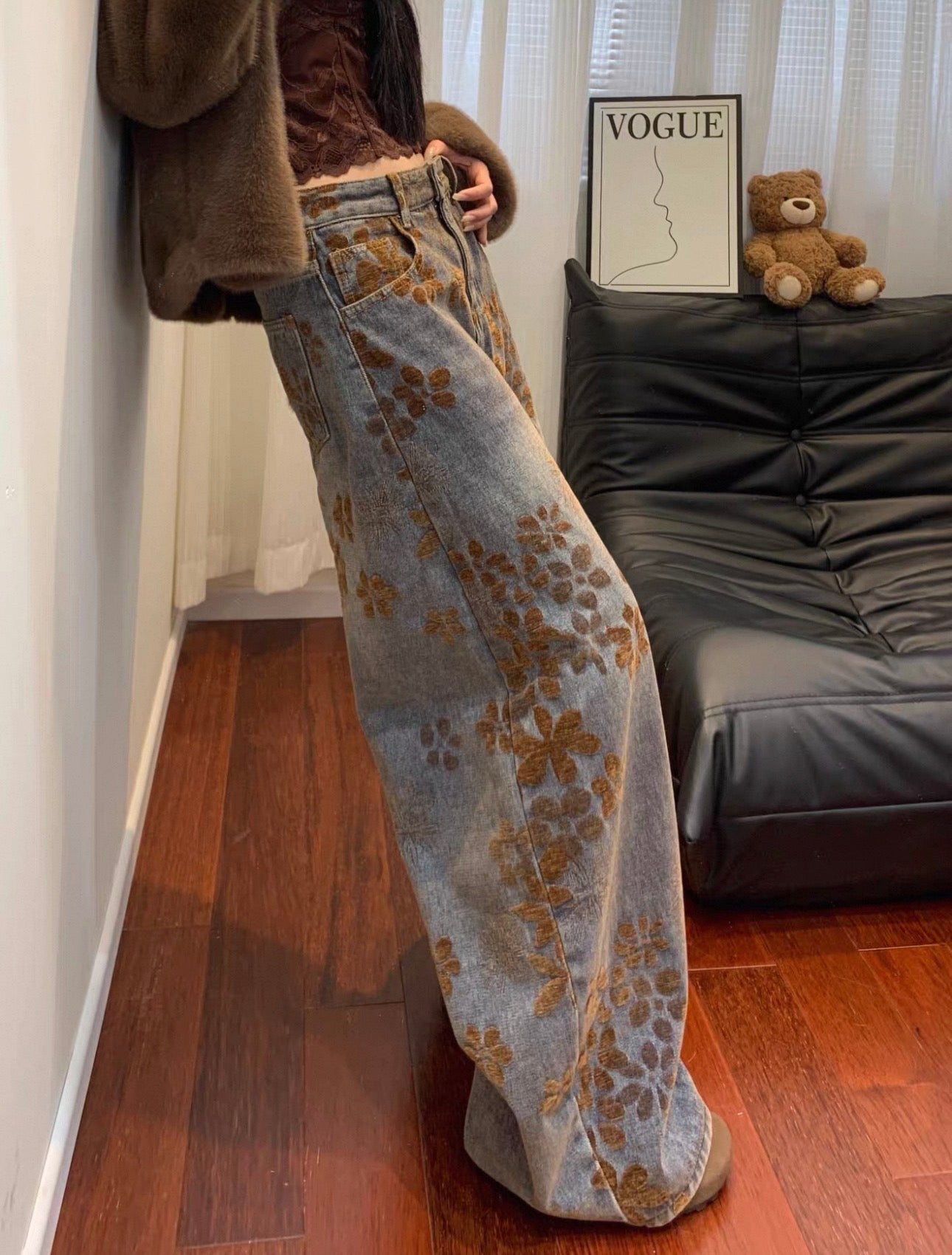 Person wearing floral-patterned jeans in a room with a black leather chair and teddy bear.