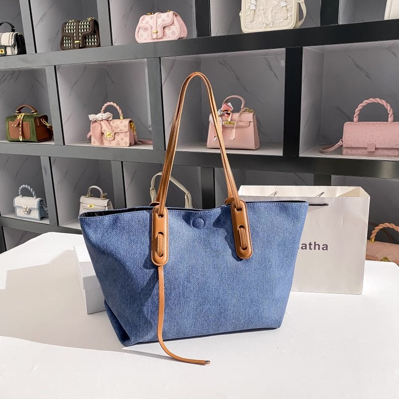 Blue tote bag with brown leather handle on a white surface, with shelves of handbags in the background.