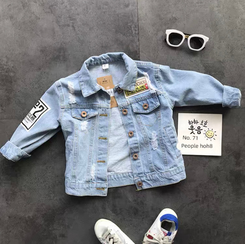 Light blue denim jacket with patches on a gray surface, accompanied by white sneakers and sunglasses.