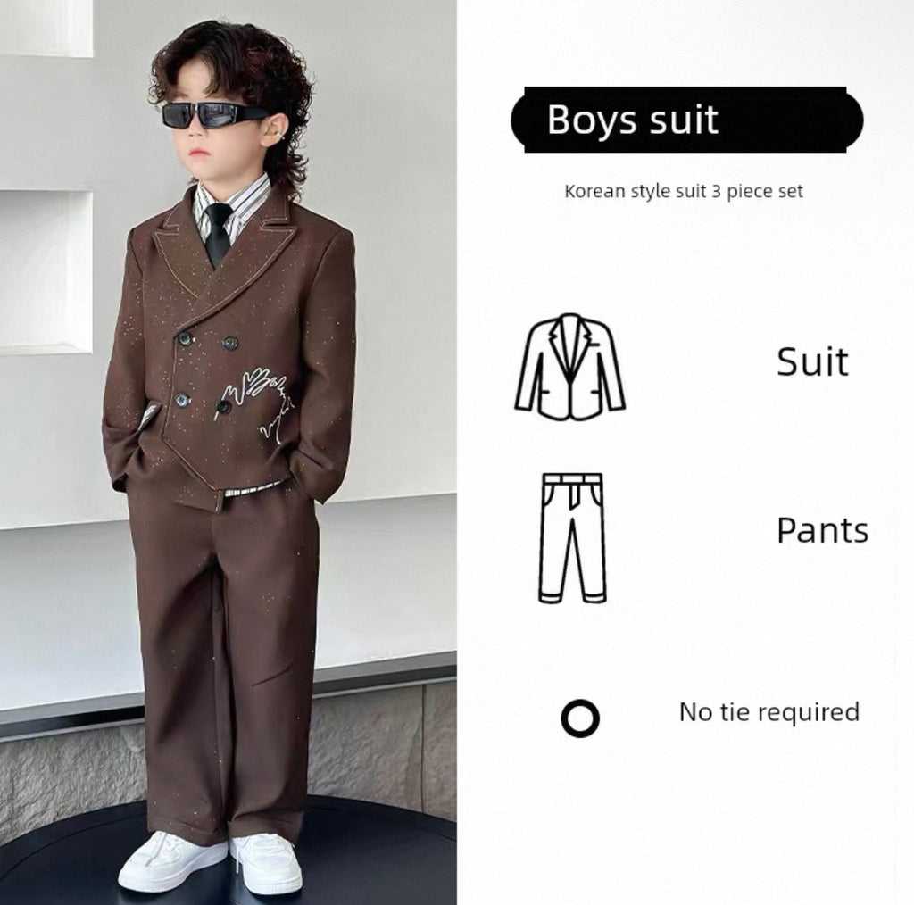 Child wearing a brown suit with a white shirt and black tie, standing against a light gray wall.