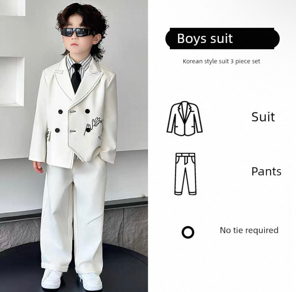 Young boy wearing a white suit with a black tie, standing in a minimalistic room.