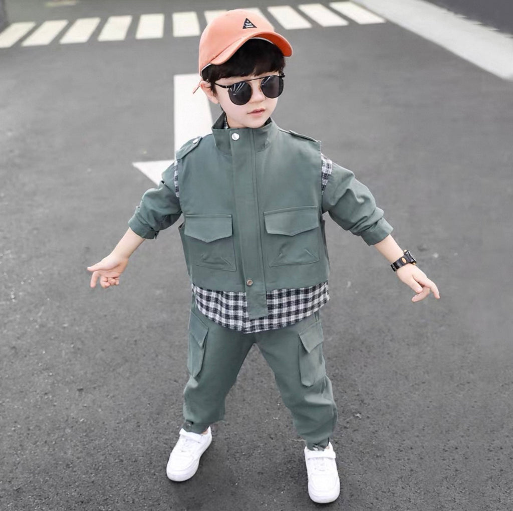 Child wearing a green outfit with a checkered shirt, white sneakers, and a cap on an asphalt surface.