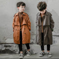 Two children wearing long coats standing against a textured wall.