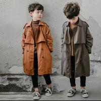 Two children wearing long coats standing against a textured wall.