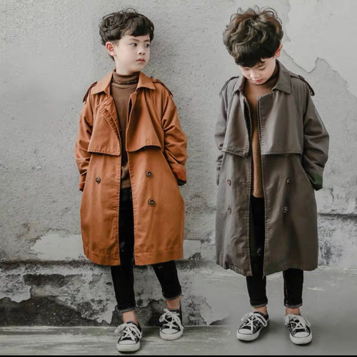 Two children wearing long coats standing against a textured wall.