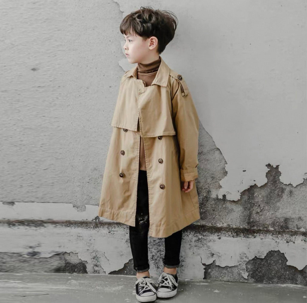 Child wearing a beige trench coat standing against a textured wall.