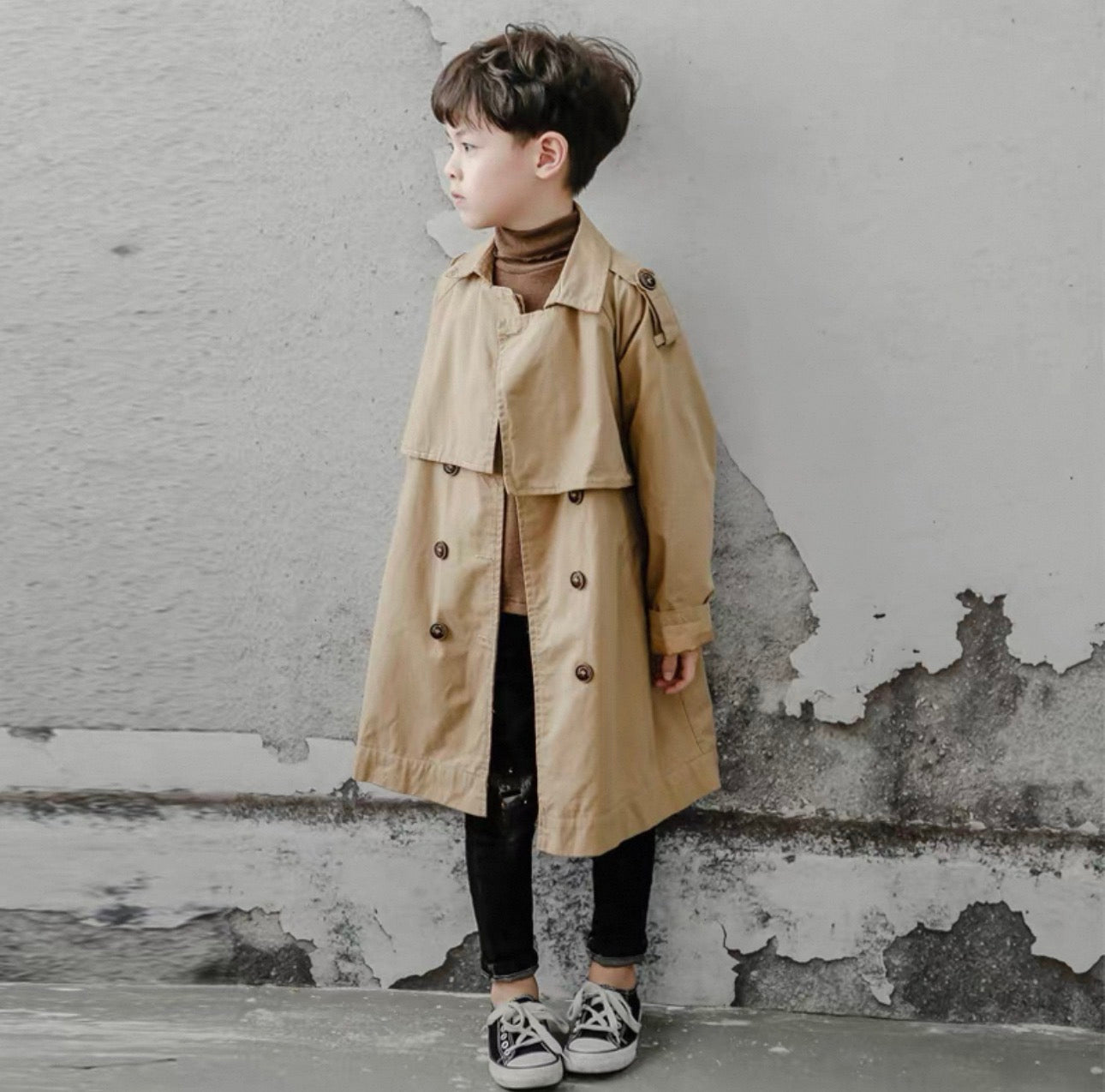 Child wearing a beige trench coat standing against a textured wall.