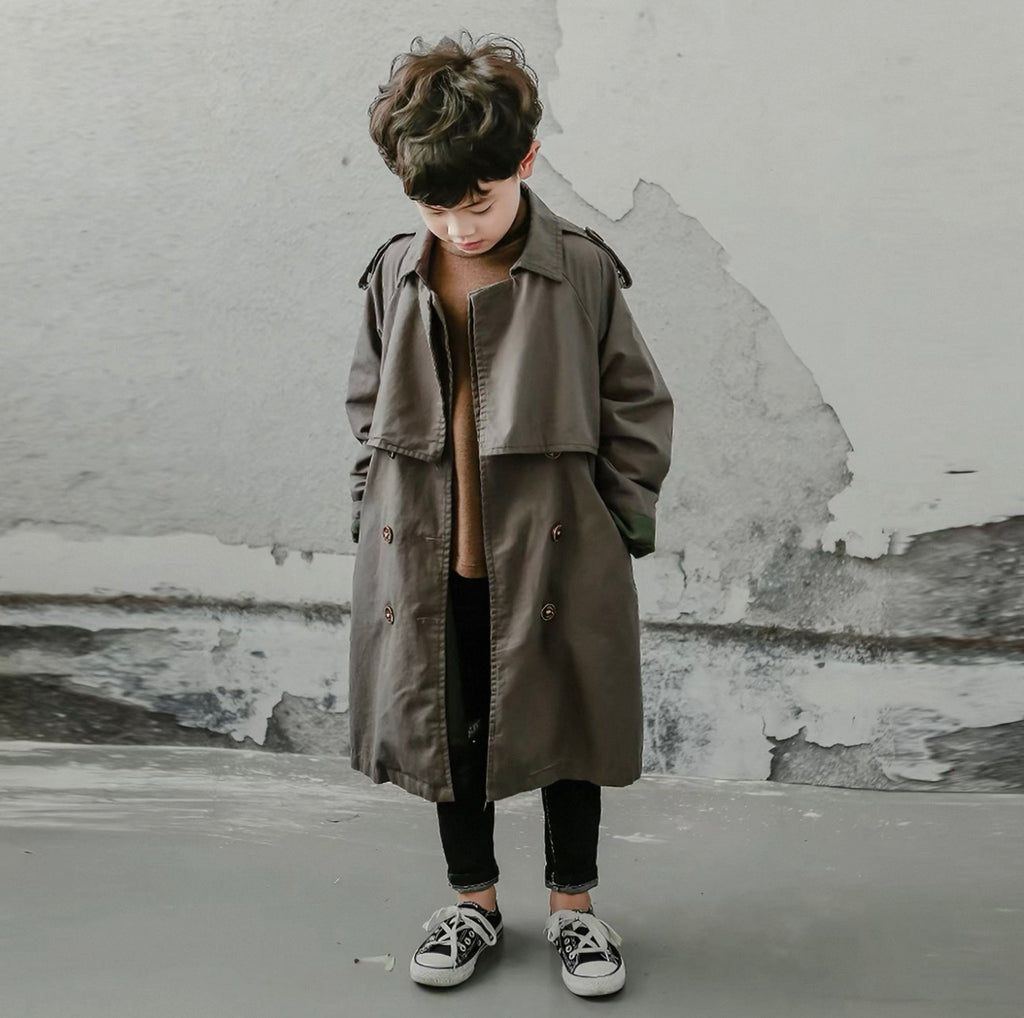 Child wearing a long charcoal coat standing against a textured wall.