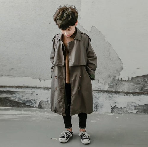 Child wearing a long charcoal coat standing against a textured wall.