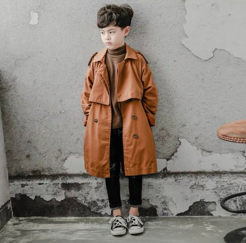 Child wearing a brown trench coat standing against a textured wall.