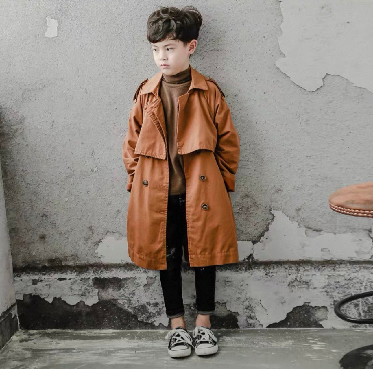 Child wearing a brown trench coat standing against a textured wall.