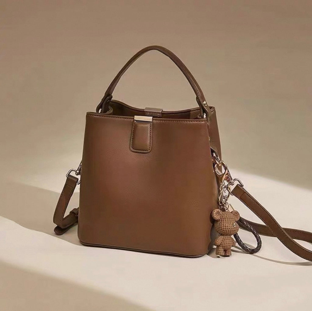 Brown leather bucket bag with single top handle and detachable shoulder strap, adorned with small teddy bear charm.