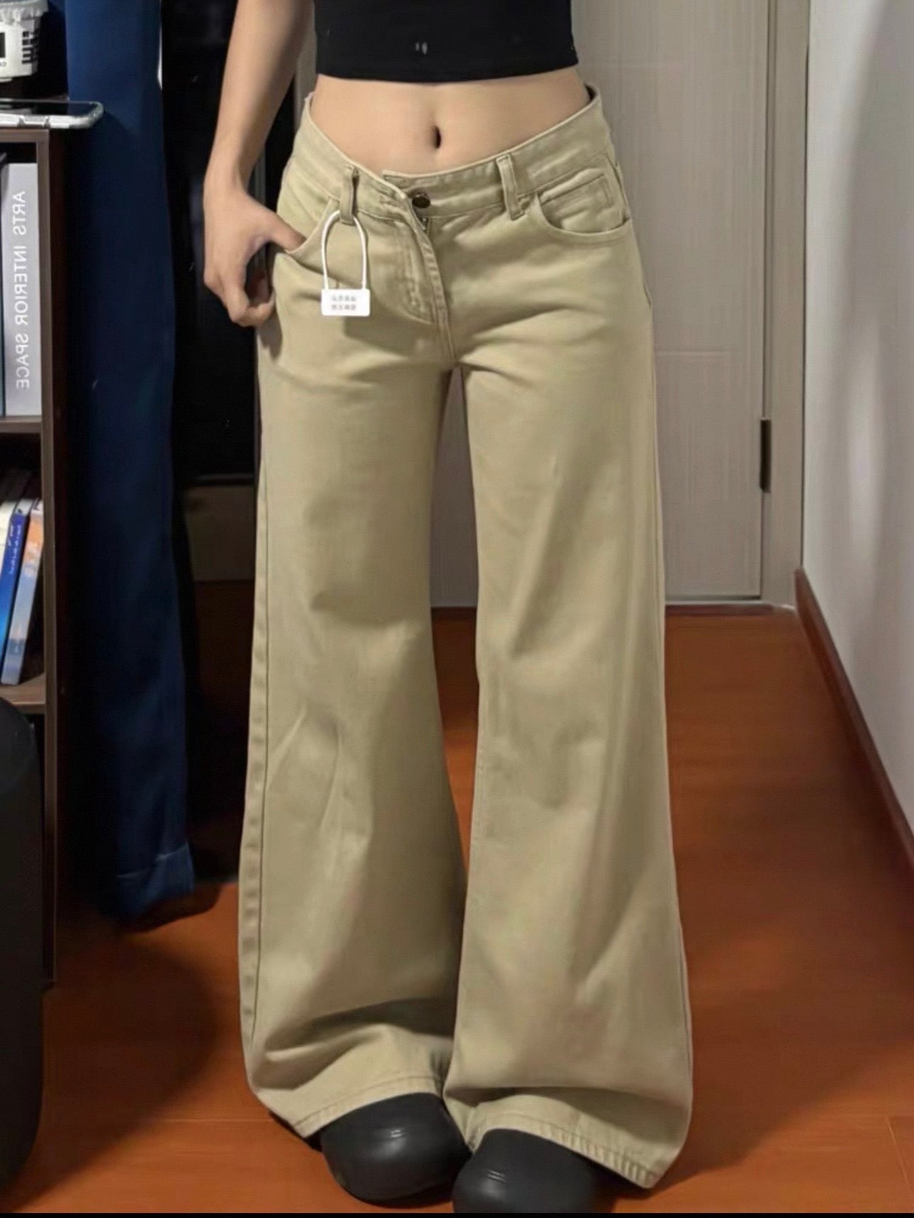Person wearing beige wide-leg pants with a clothing tag, standing indoors.