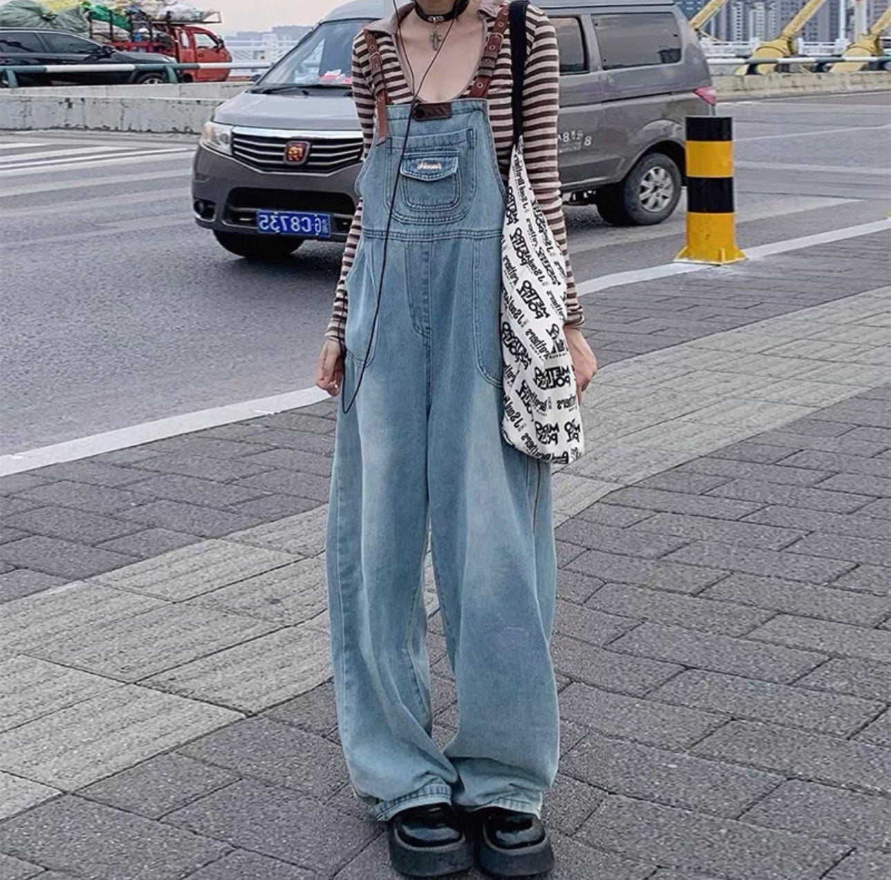 Person wearing a striped shirt and denim overalls standing on a street.