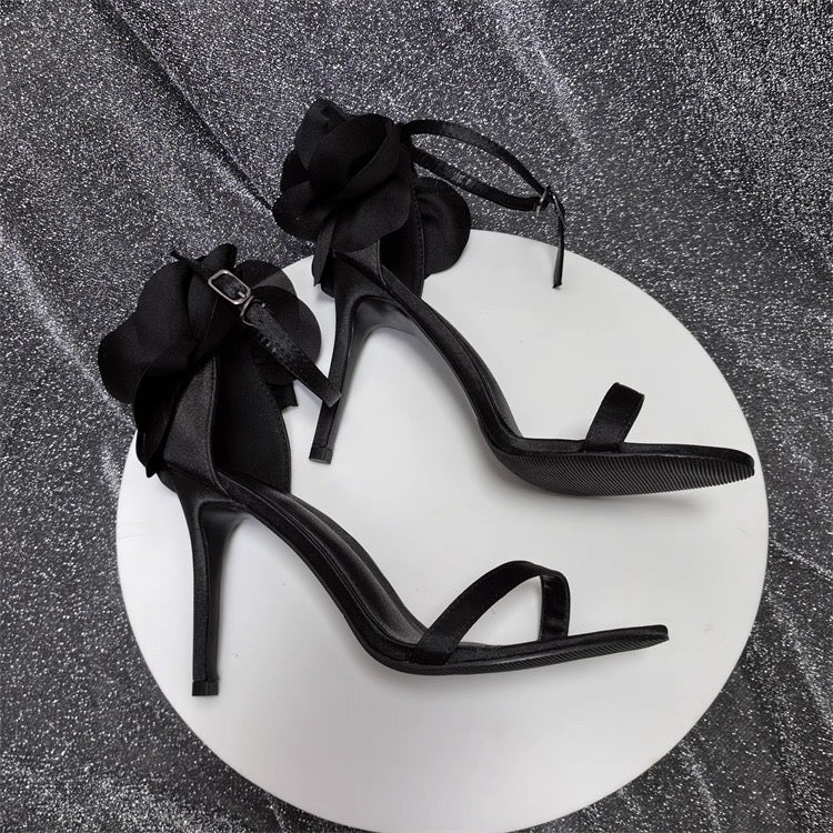 Black satin stiletto sandals with ankle strap and large floral embellishment on heel.