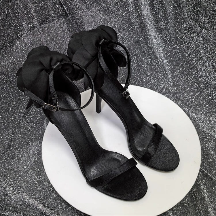 Black satin stiletto sandals with ankle strap and floral ruffle detail on back.