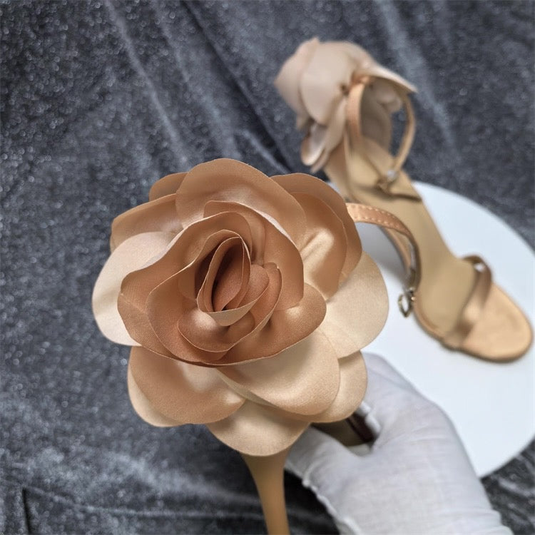 Nude satin stiletto sandals featuring a large floral embellishment on the ankle strap.