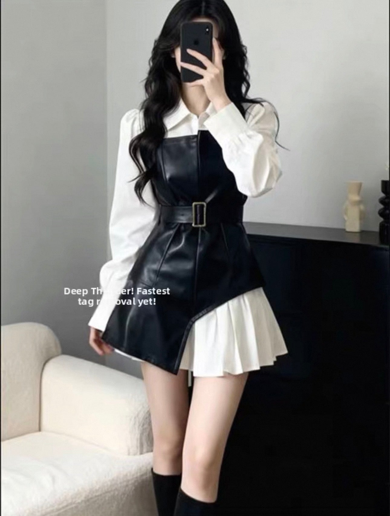 Person wearing a black leather dress over a white shirt with a pleated skirt, sitting on a couch.