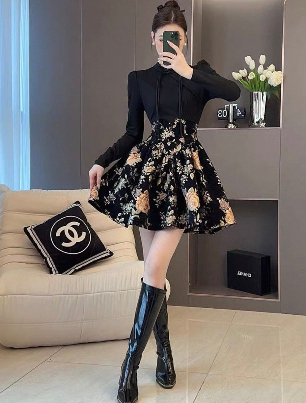 Person wearing a black long-sleeve top and floral skirt with knee-high black boots in a living room.