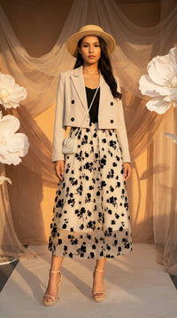 Woman in a stylish outfit with a floral skirt and beige jacket standing against a decorative backdrop.