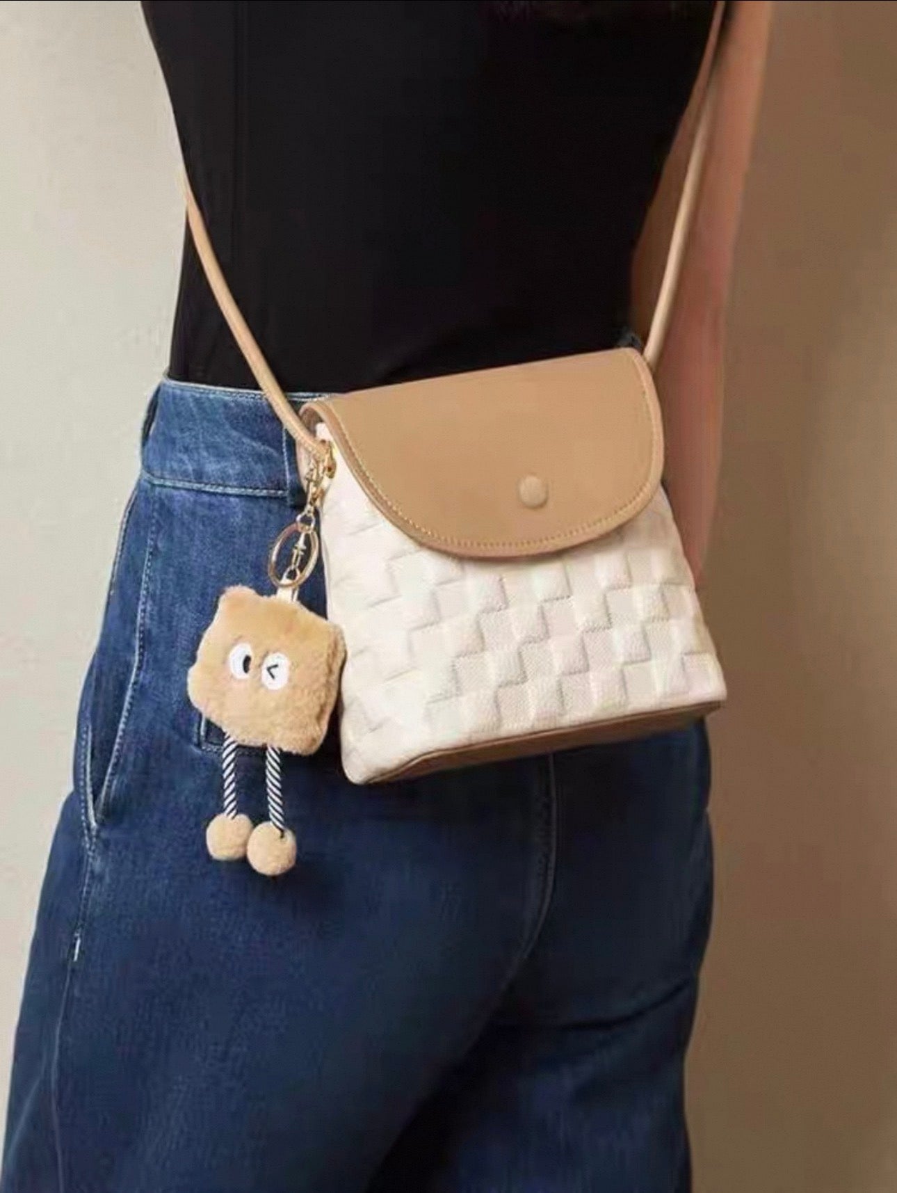 Beige small square crossbody bag with woven texture and flap closure, paired with a cute plush keychain charm.