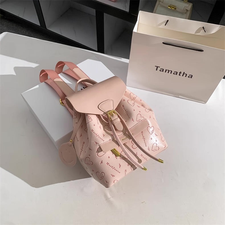 Pink backpack with heart patterns on a white surface, next to a 'Tamatha' branded box.