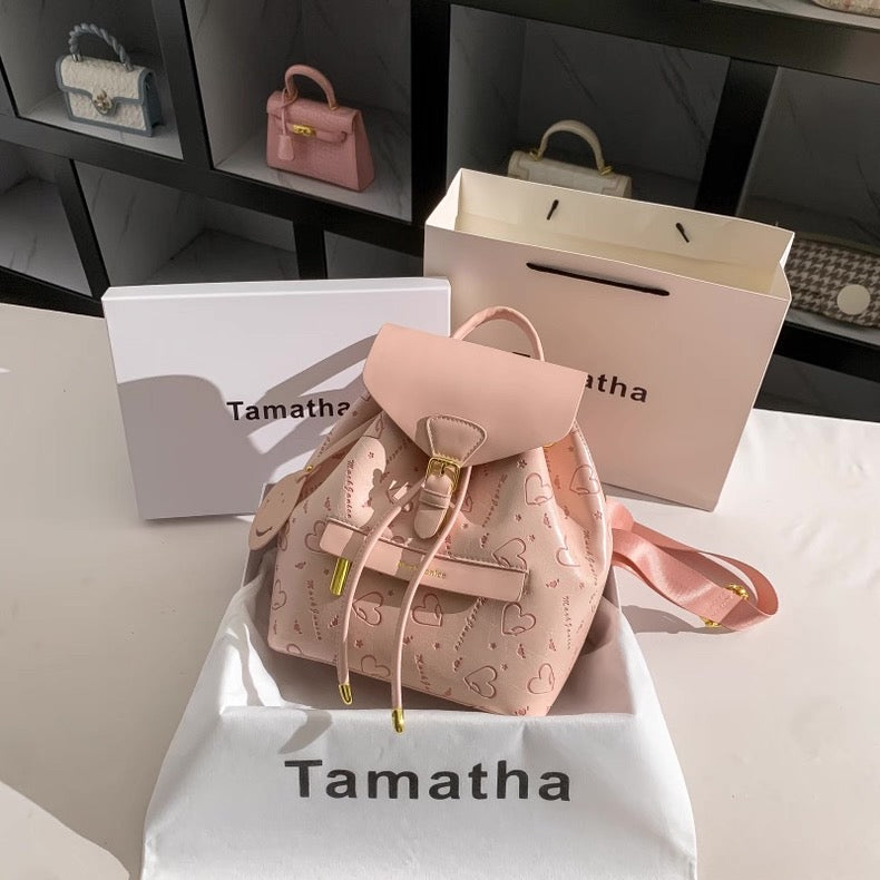 Pink backpack with heart patterns on a white box labeled 'Tamatha'.