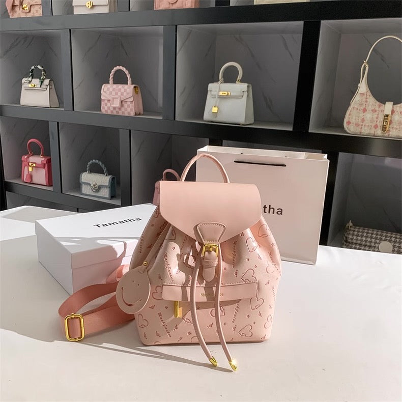 Pink backpack with heart patterns on a white surface, with shelves of handbags in the background.