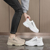 Person holding a white sneaker with zippers next to beige and white sneakers on a wooden floor.