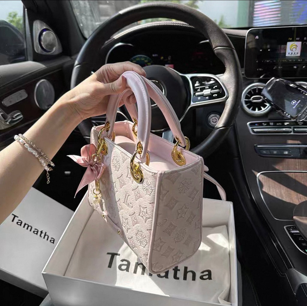 Light pink embossed crossbody bag with gold hardware and decorative charm, held inside a car interior.