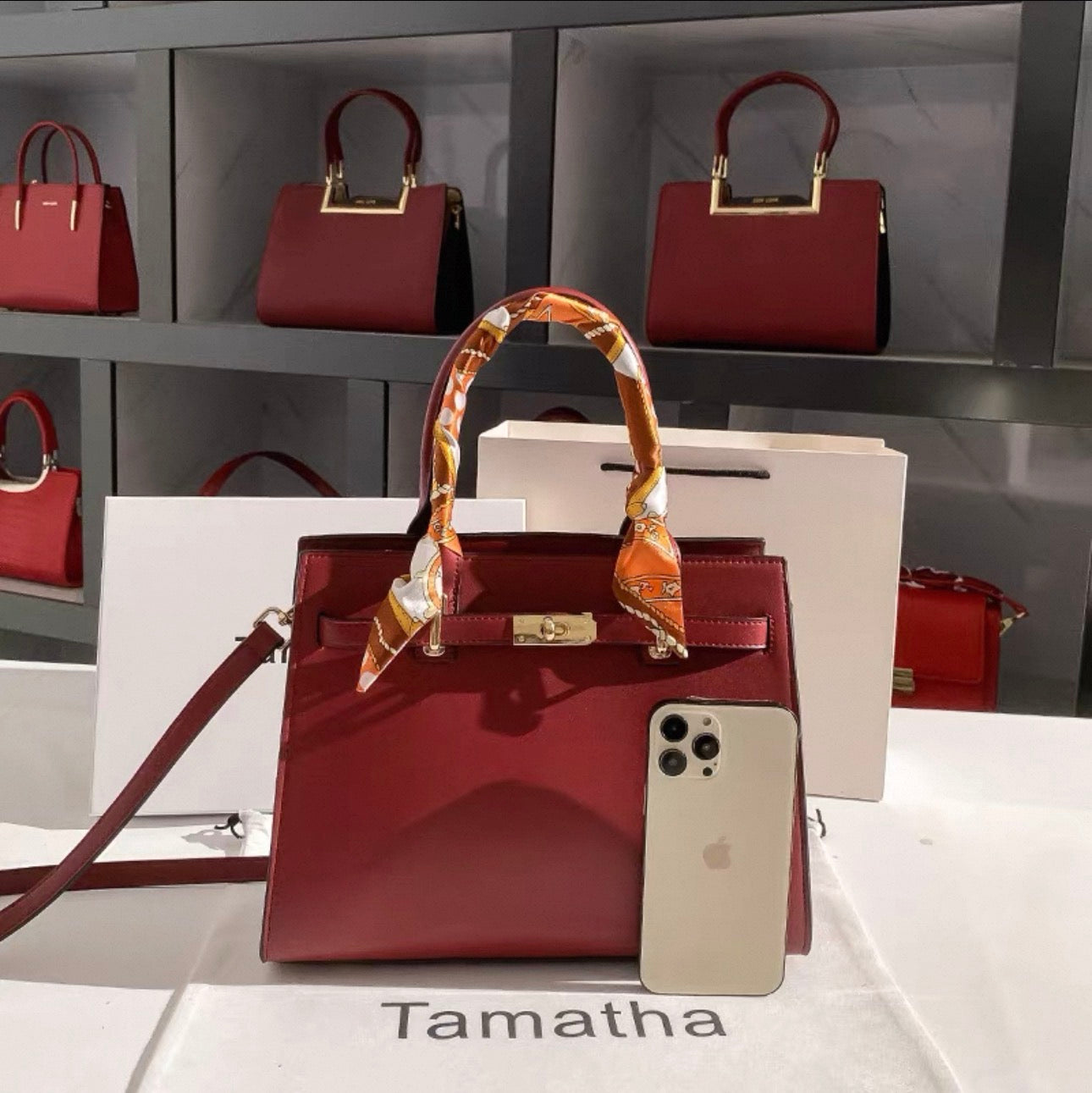 Red high-end Kelly-style handbag with glossy finish and a patterned silk scarf wrapped around the handle, displayed with an.