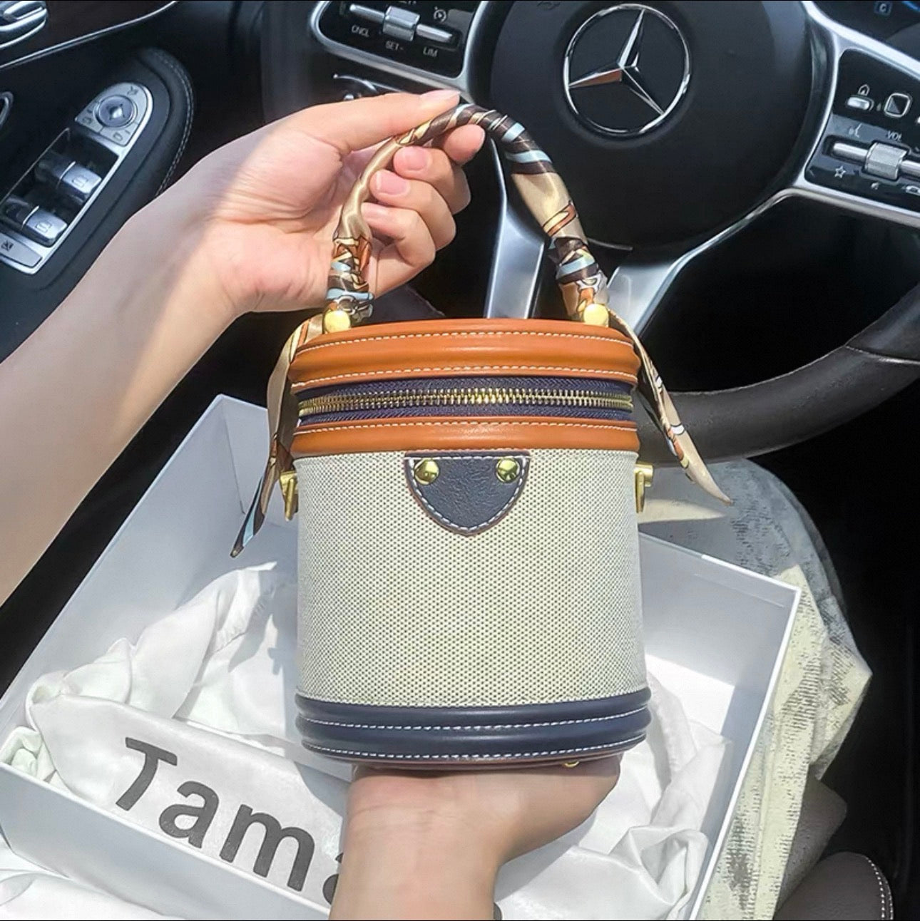Multicolor cylindrical crossbody bag with beige canvas body, dark brown and orange leather accents, and bamboo-style top.
