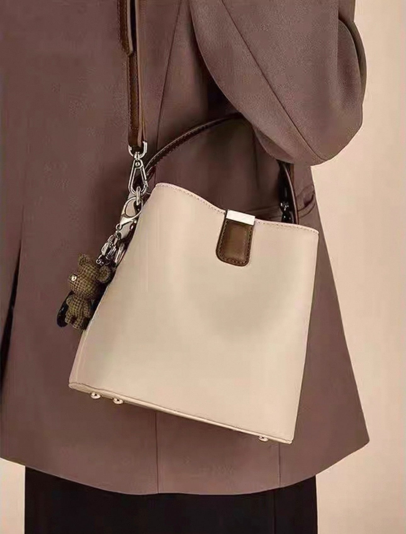 Beige bucket bag with single shoulder strap and silver hardware worn over brown blazer.