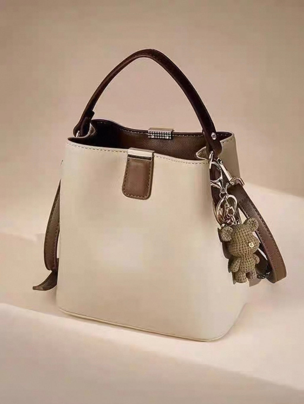 Beige bucket handbag with single shoulder strap and teddy bear charm attached.