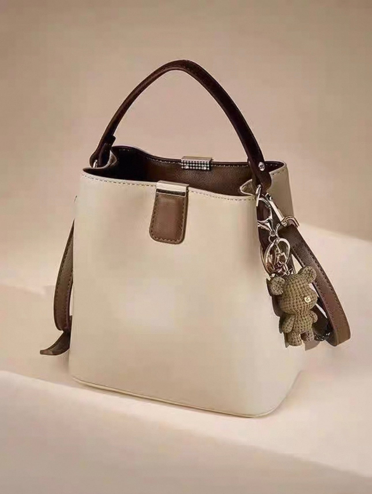 Beige bucket handbag with single shoulder strap and teddy bear charm attached.