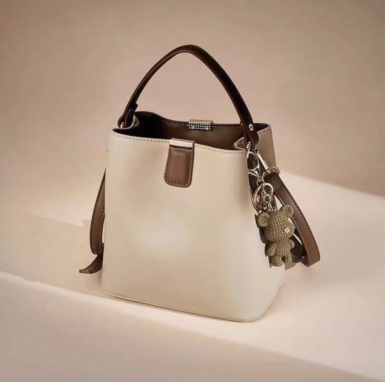 Beige bucket bag with brown handle, silver hardware, and teddy bear keychain charm.