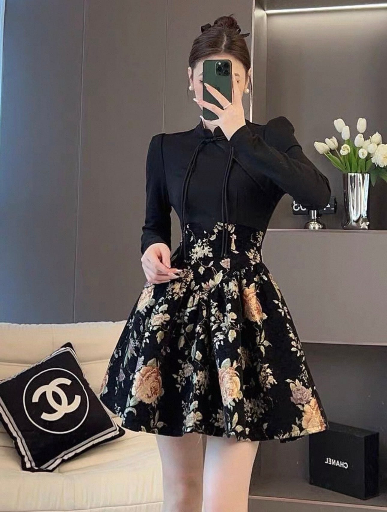 Person wearing a black long-sleeve top and floral skirt in a room with a sofa and vase.