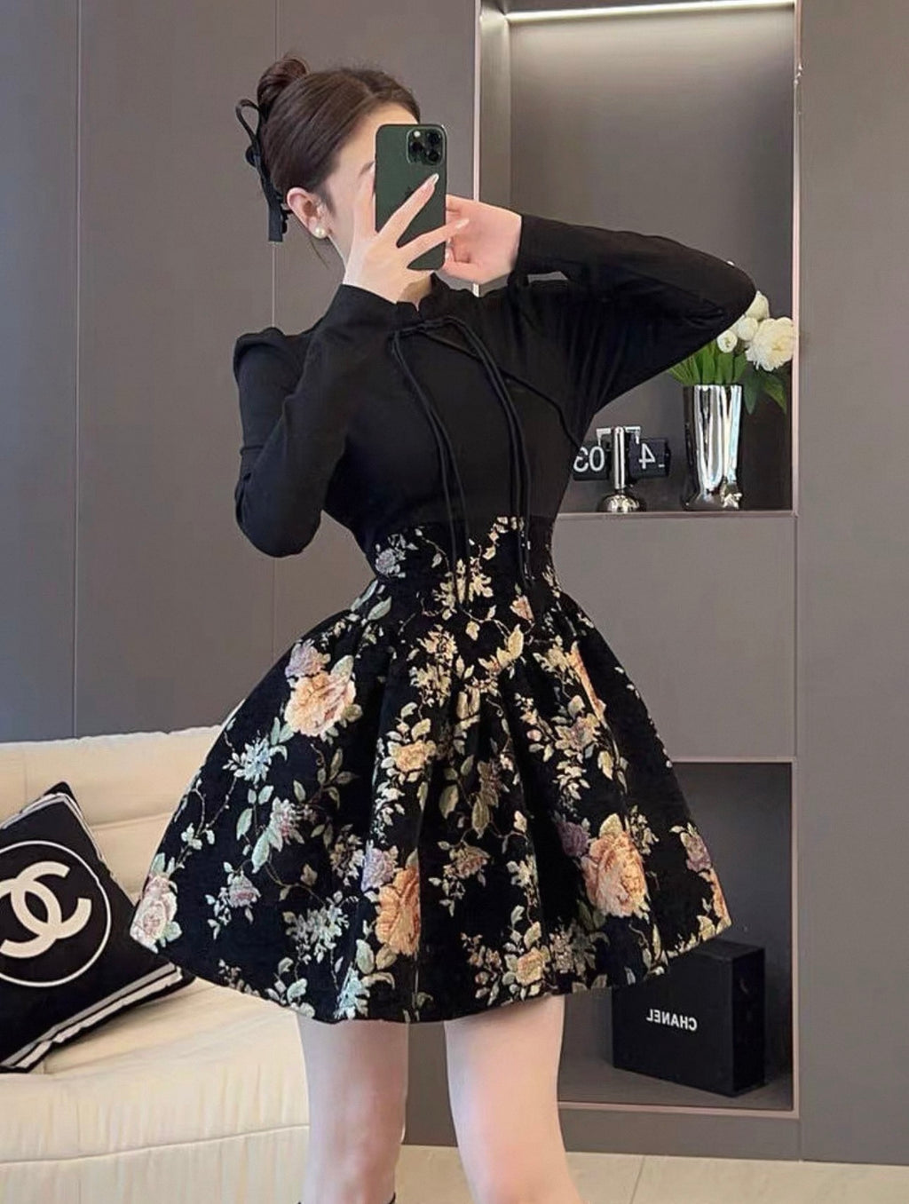 Woman taking a mirror selfie wearing a black long-sleeve top and floral skirt in a room.