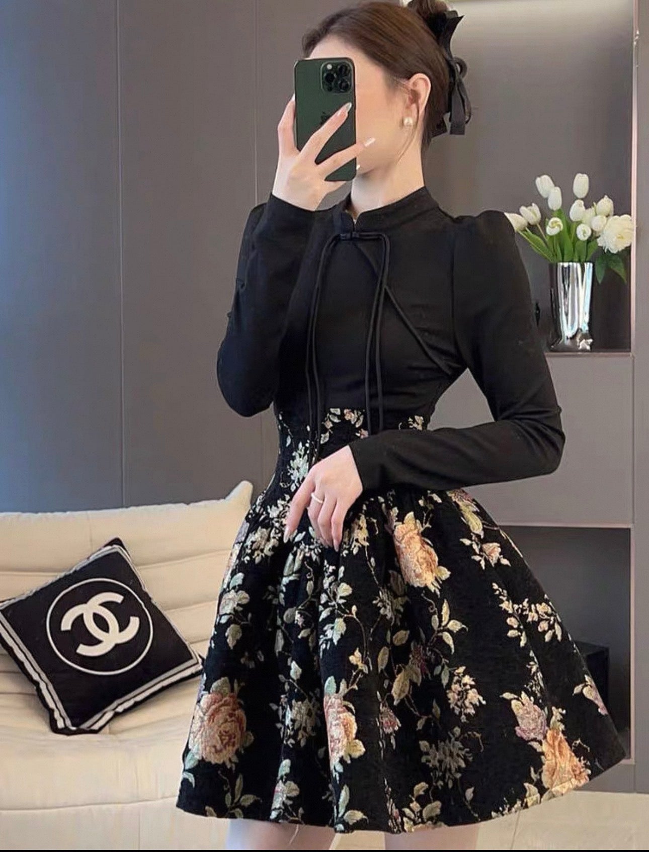 Person wearing a black long-sleeve top and floral skirt in a room with a couch and vase.