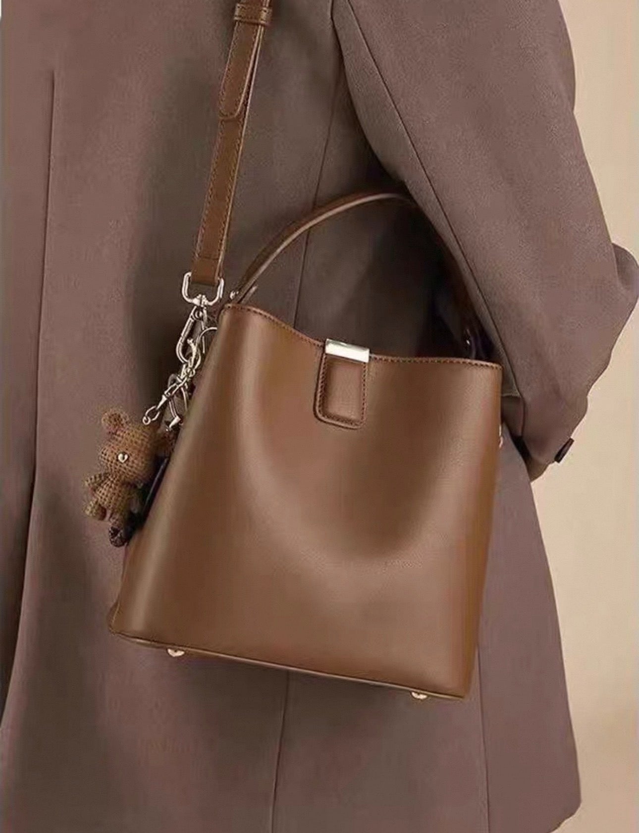 Brown leather bucket bag with single top handle and detachable shoulder strap, silver hardware and teddy bear charm hanging.